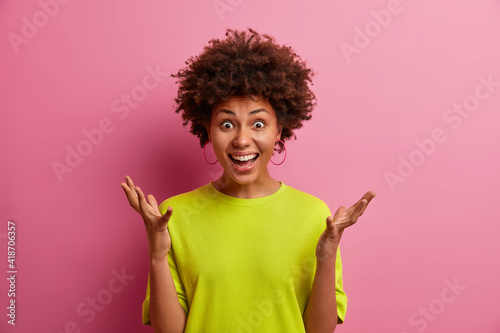 Human reaction concept. Cheerful beautiful young Afro American woman raises hands reacts positively on great news dressed in casual clothes isolated over pink background. Wow how great it is