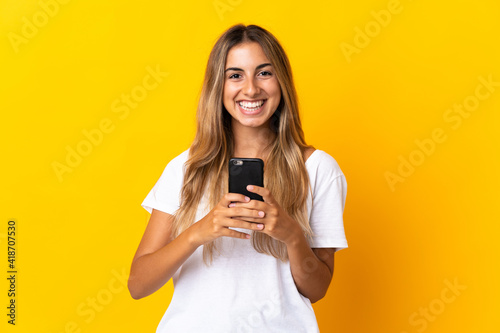 Young hispanic woman over isolated yellow background looking at the camera and smiling while using the mobile
