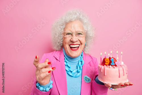 Life only starts when get older. Amused fashionable senior lady celebrates birthday holds cake with burning candles wears makeup isolated over pink background. Positive granny celebrates 91st bday