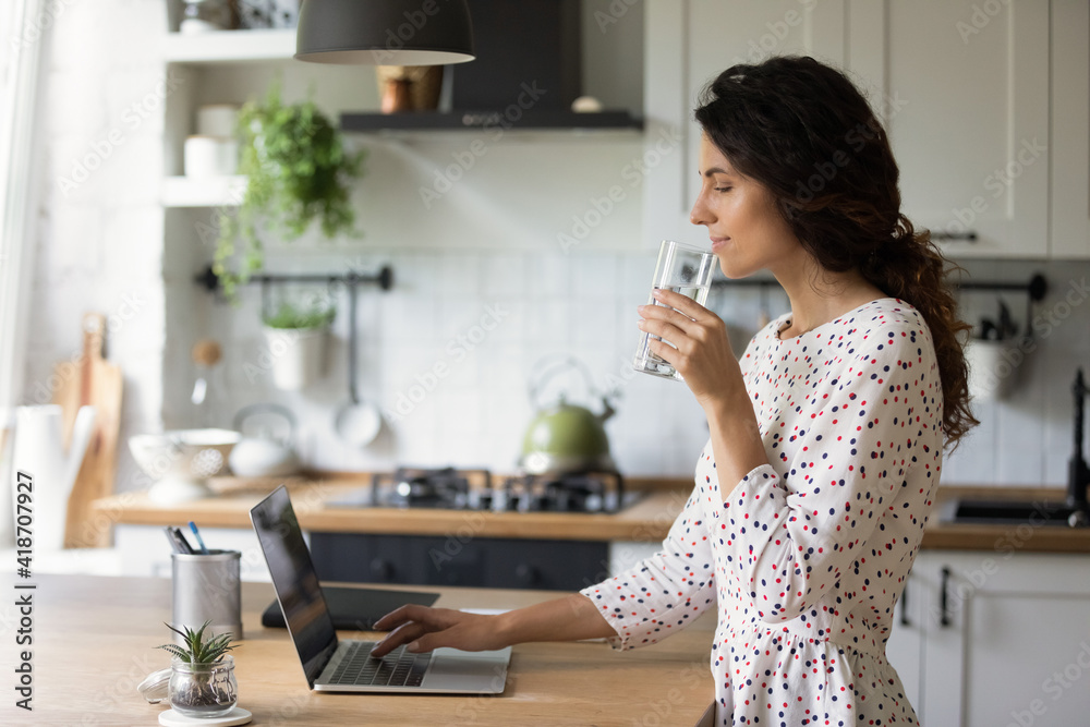 Healthy habit. Relaxed female freelancer working from home start work day from glass of pure crystal water check mail read news on laptop. Happy young woman drink mineral aqua at kitchen by pc screen. Healthy habit. Relaxed female freelancer working from home start work day from glass of pure crystal water check mail read news on laptop. Happy young woman drink mineral aqua at kitchen by pc screen.