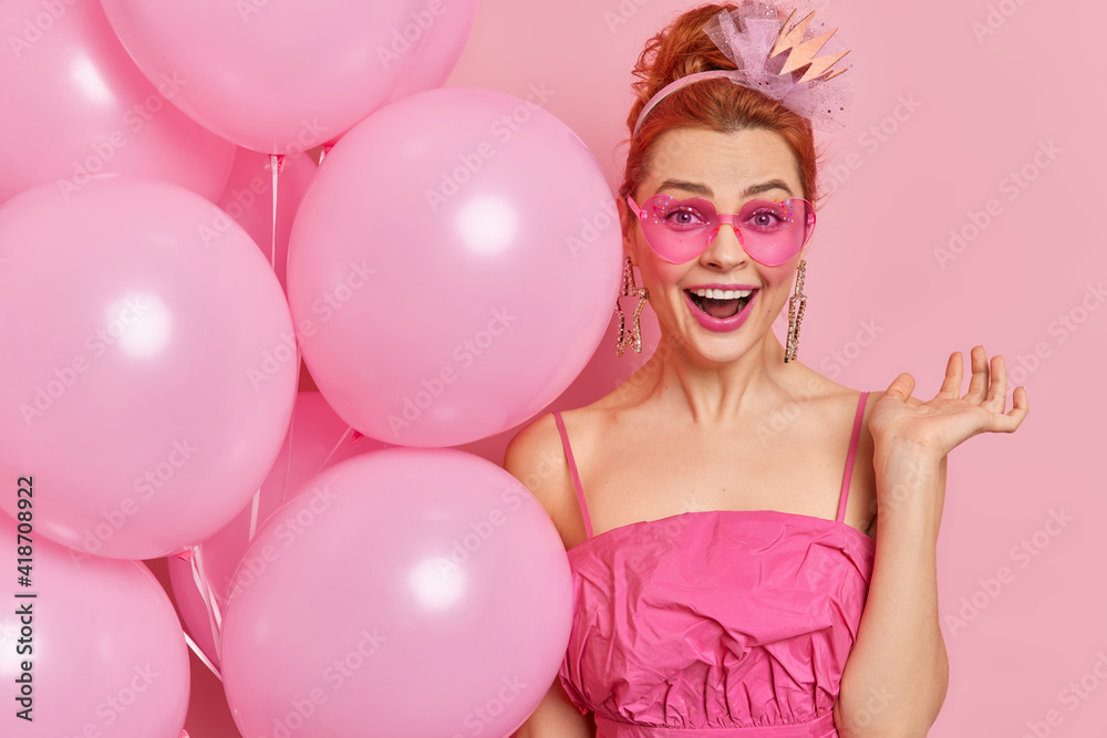 Happy redhead young woman wears trendy heart shaped sunglasses and dress holds bunch of inflated balloons celebrates birthday has positive mood isolated over pink background. Festivity concept
