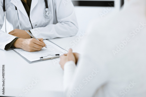 Unknown woman-doctor and female patient sitting and talking at medical examination in clinic  close-up. Therapist wearing green blouse is filling up medication history record. Medicine concept