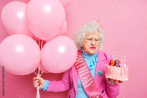 Unhappy elderly beautiful woman sad about getting older looks at delicious cake with burning candles celebrates 91st birthday hollds bunch of balloons accepts congratulations on party. Aging concept © wayhome.studio 