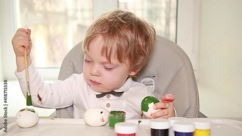 A little boy in a white shirt paints chicken eggs with paints. Preparations for coloring Easter eggs. Preparing for Easter. Learning and creative development for children. Selective focus A little boy in a white shirt paints chicken eggs with paints. Preparations for coloring Easter eggs. Preparing for Easter. Learning and creative development for children. Selective focus