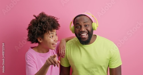 Hey look there. Positive Afro Americann woman points directly at camera shows something funny to boyfriend who listens audio track via headphones giggle together stands closely indoor over pink wall photo