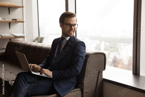 Smiling businessman wearing glasses and suit distracted from laptop, sitting on couch in office, looking to aside, successful entrepreneur visualizing good future, dreaming about new opportunities © fizkes