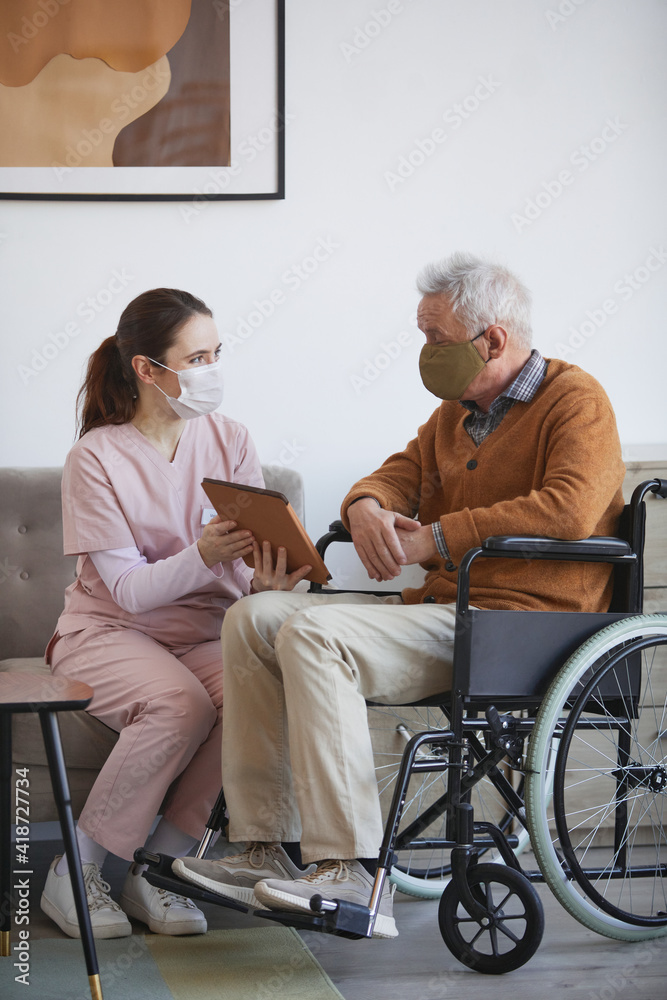 Vertical full length portrait of young female nurse assisting senior man in wheelchair using digital tablet at retirement home, both wearing masks Vertical full length portrait of young female nurse assisting senior man in wheelchair using digital tablet at retirement home, both wearing masks
