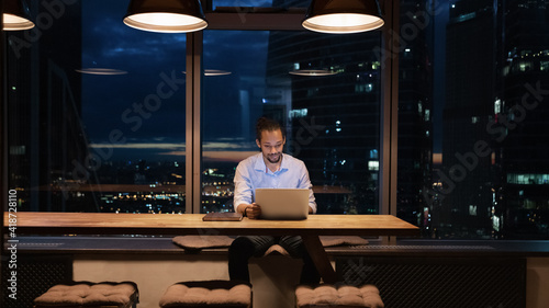 Full length African American businessman working on laptop in office at night alone  looking at screen  sitting at desk near panoramic window  employee finishing project at late hours  deadline