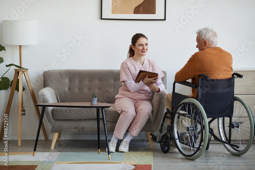 Full length portrait of smiling female nurse talking to senior man in wheelchair and using digital tablet at retirement home, copy space photo
