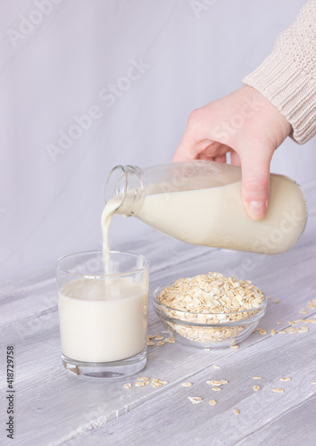 A hand pours vegetable oat milk from a bottle into a glass tumbler on a light background. Healthy drinks, vegetarianism. Side view. Vertical photo