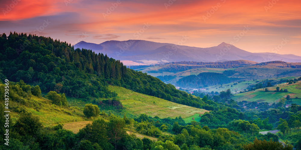 mountainous rural landscape at dawn. beautiful scenery with forests, hills and meadows in morning light. ridge with high peak in the distance. village in the distant valley