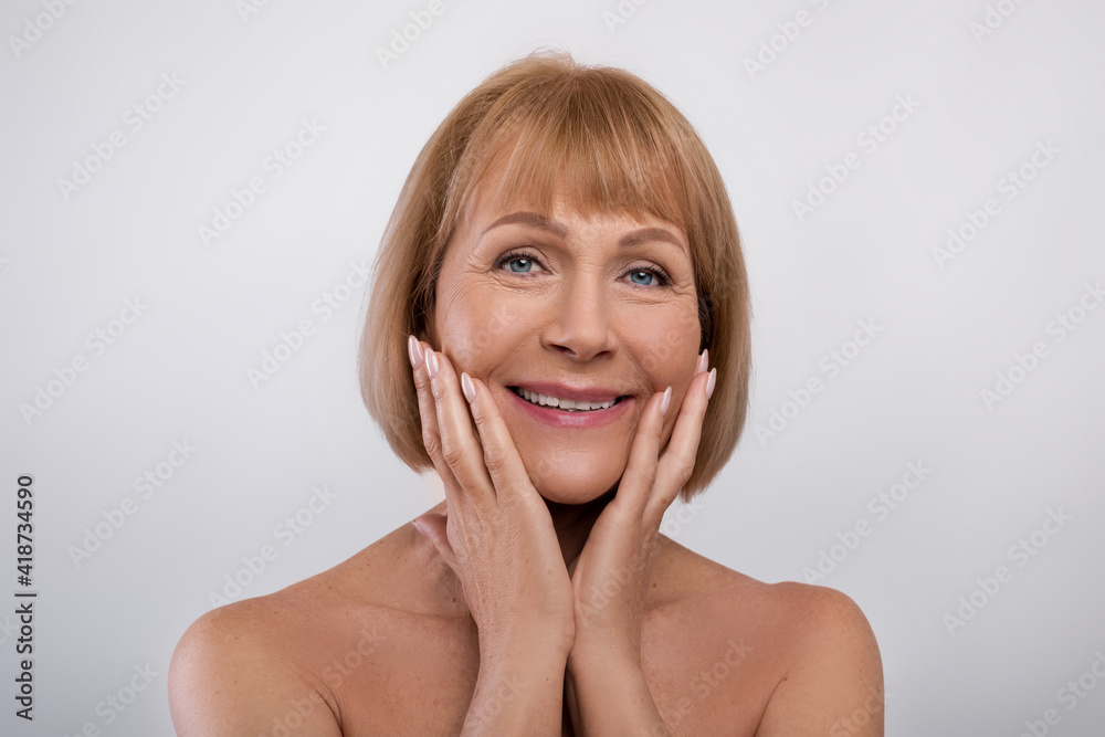 Lovely senior lady with perfect skin touching her face and smiling at camera over blue studio background Lovely senior lady with perfect skin touching her face and smiling at camera over blue studio background