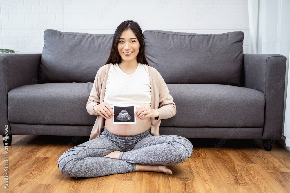 beautiful asian pregnant woman sitting on sofa holding ultrasound baby picture and smiling with happiness, resting relaxing in living room from hormone stress, wearing comfy stretch pants and jumper