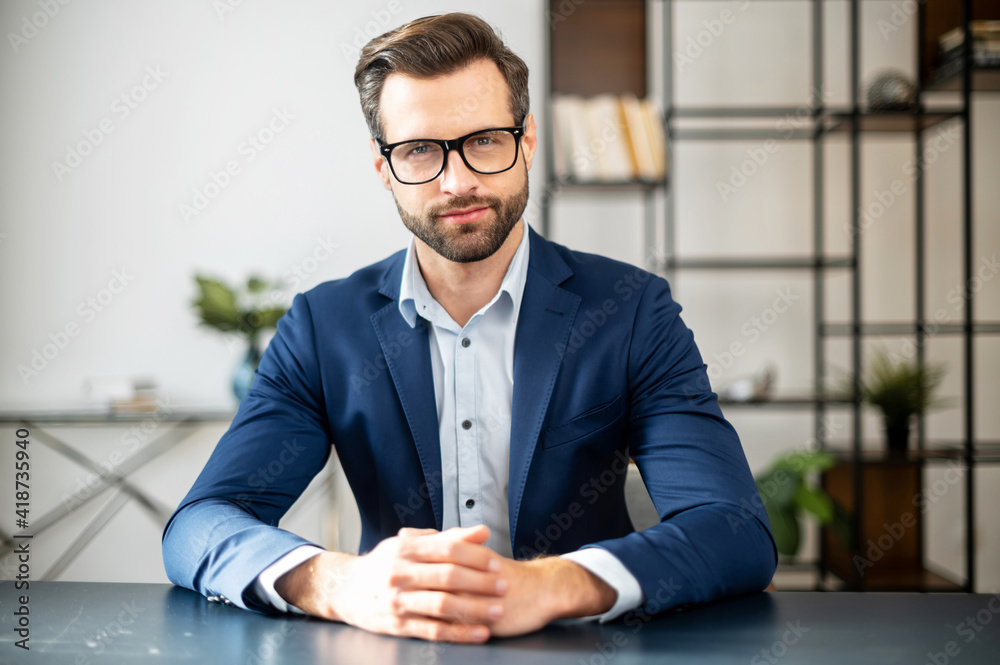 Successful handsome businessman sitting at desk, talking at camera, sharing experience and ideas, free space, time to be productive, in tech start-up office, employer lead job interview with applicant Successful handsome businessman sitting at desk, talking at camera, sharing experience and ideas, free space, time to be productive, in tech start-up office, employer lead job interview with applicant