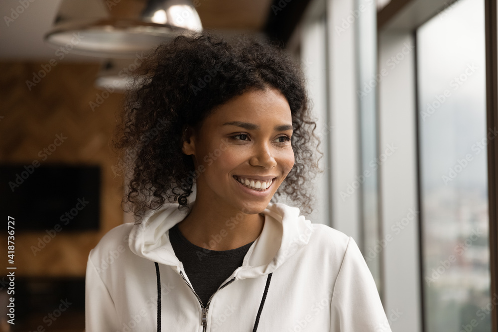 Head shot smiling dreamy African American young woman looking to aside, standing at home near window, thinking, visualizing good future, dreaming about new opportunities, planning, enjoying view Head shot smiling dreamy African American young woman looking to aside, standing at home near window, thinking, visualizing good future, dreaming about new opportunities, planning, enjoying view