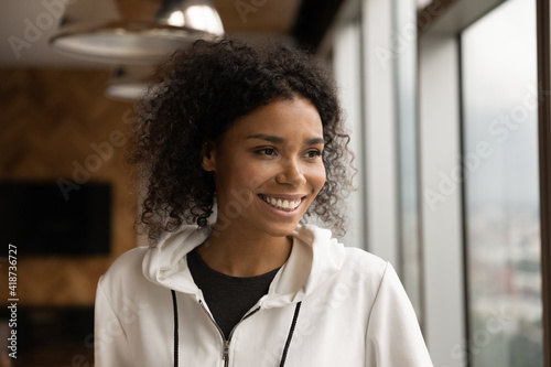 Head shot smiling dreamy African American young woman looking to aside, standing at home near window, thinking, visualizing good future, dreaming about new opportunities, planning, enjoying view