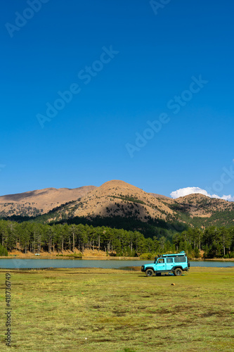 A blue SUV car parked in a meadow next to a small lake on a very high mountain top, green trees and bare mountain peaks in the background