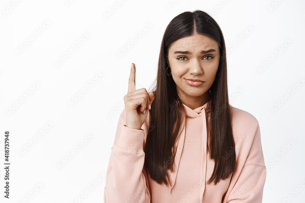Skeptical and unamused glamour girl pointing finger up, frowning and looking with disbelief and dislike, showing bad advertisement, standing over white background Skeptical and unamused glamour girl pointing finger up, frowning and looking with disbelief and dislike, showing bad advertisement, standing over white background