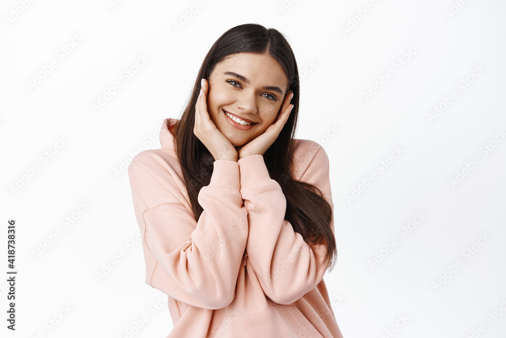 Portrait of cute and tender brunette girl blushing from happy news or surprise, holding hands on cheeks and smiling, standing against white background Portrait of cute and tender brunette girl blushing from happy news or surprise, holding hands on cheeks and smiling, standing against white background