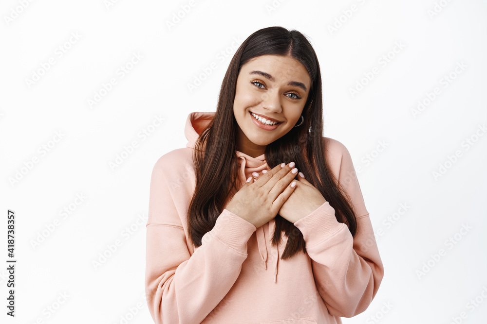 Is it for me. Cute heartfelt girl receive a gift, holding hands on heart and smiling pleased, say thank you, express gratitude, standing in hoodie against white background Is it for me. Cute heartfelt girl receive a gift, holding hands on heart and smiling pleased, say thank you, express gratitude, standing in hoodie against white background