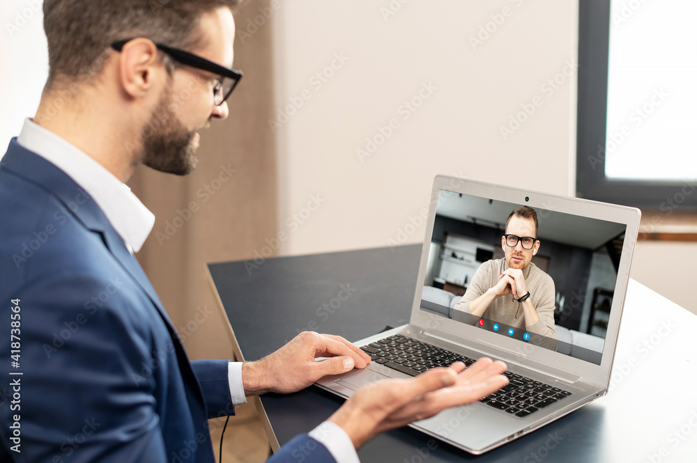 Successful employee with glasses in elegant suit, sitting at the laptop, online business meeting with potential clients and partners, looking at the laptop, working remotely