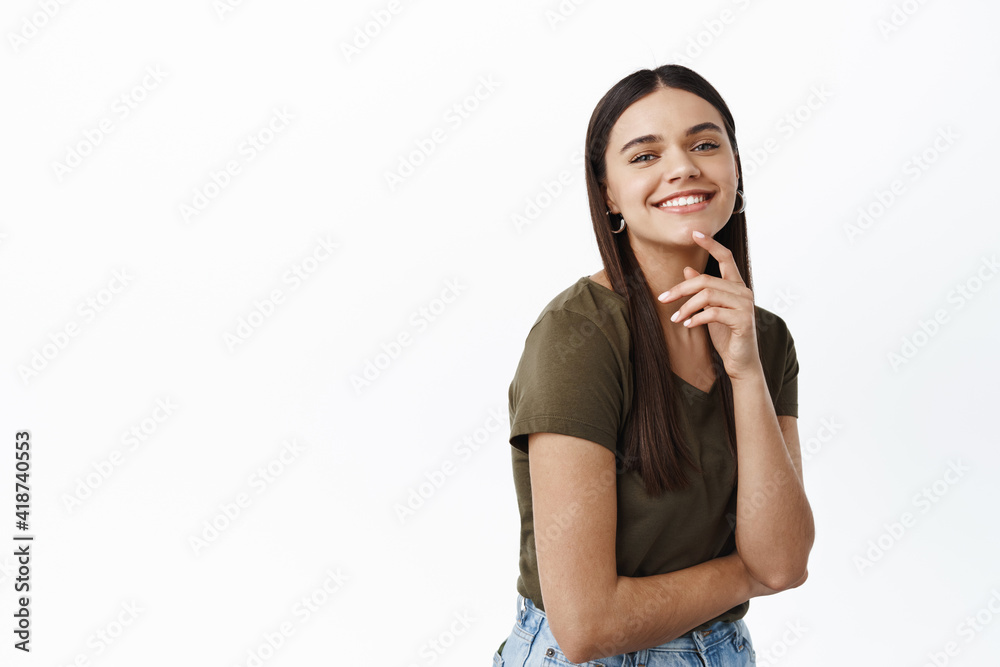 Portrait of young successful and happy woman smiling at camera, express joy and positive emotions, touching natural clean face, standing against white background Portrait of young successful and happy woman smiling at camera, express joy and positive emotions, touching natural clean face, standing against white background