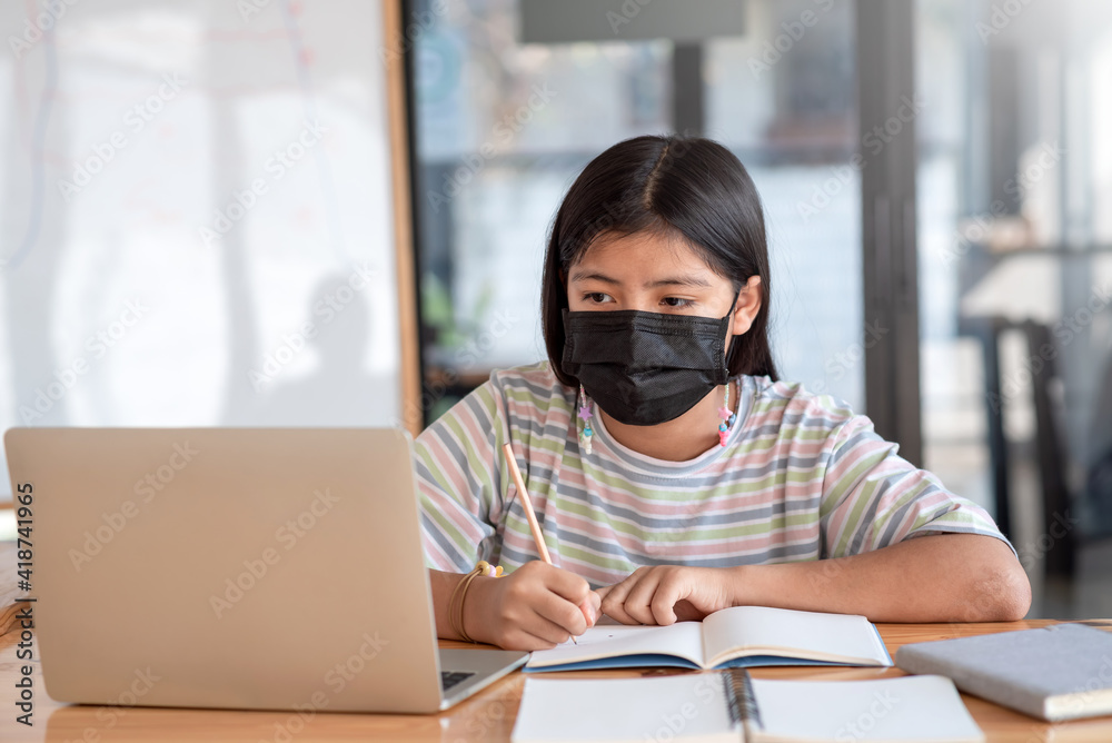 Asian young girl sitting at the table Wear a mask To prevent germs, take notes, look at the laptop at home. Asian young girl sitting at the table Wear a mask To prevent germs, take notes, look at the laptop at home.