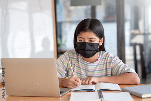 Asian young girl sitting at the table Wear a mask To prevent germs, take notes, look at the laptop at home.