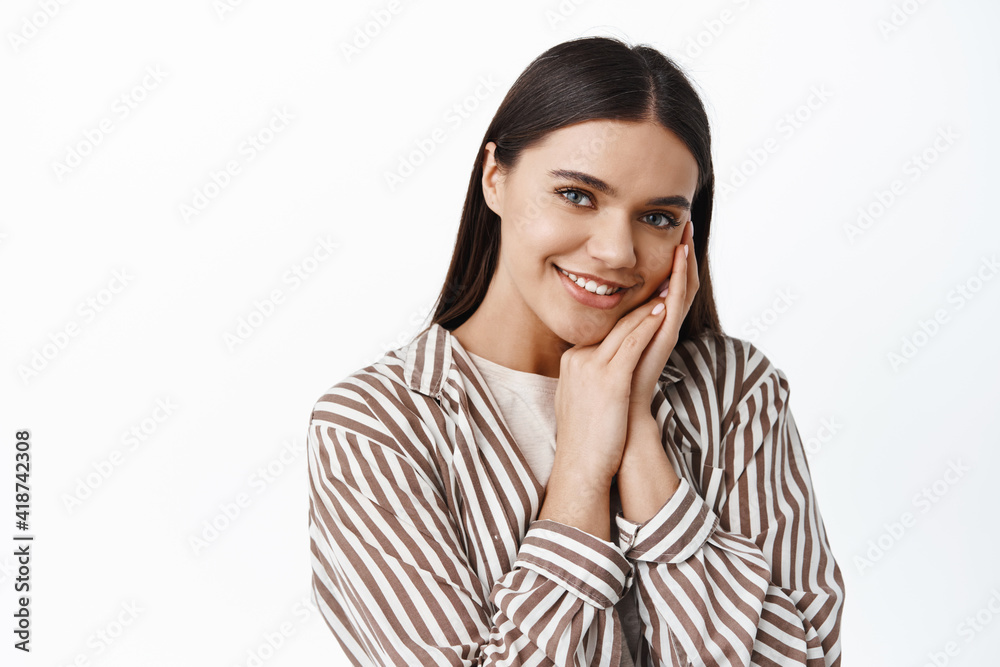 Close up portrait of beautiful smiling woman, gently touching natural clean skin face, gazing at camera delighted, standing against white background Close up portrait of beautiful smiling woman, gently touching natural clean skin face, gazing at camera delighted, standing against white background