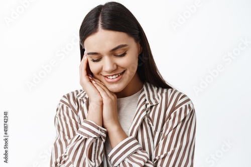 Close up portrait of tender and feminine young woman, looking down and touching soft clean skin with natural light make up, standing over white background