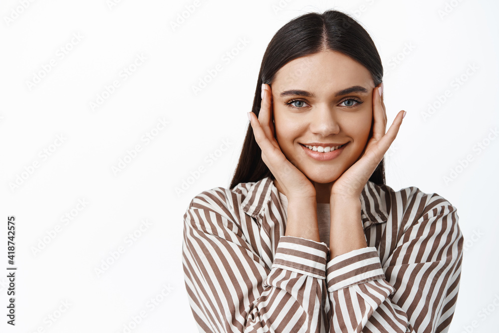 Beautiful brunette girl with happy smile, touching face with fingertips near eyes, using skin care routing cosmetics for nourished facial result, white background Beautiful brunette girl with happy smile, touching face with fingertips near eyes, using skin care routing cosmetics for nourished facial result, white background