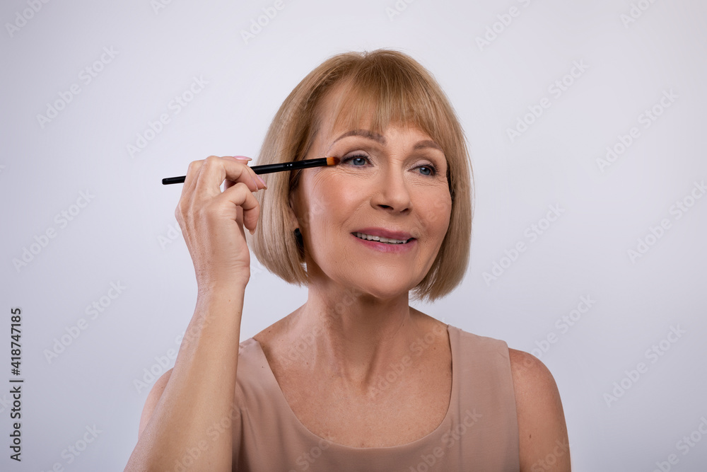Portrait of lovely senior woman applying eye shadow on her face over light studio background. Beauty and wellness Portrait of lovely senior woman applying eye shadow on her face over light studio background. Beauty and wellness