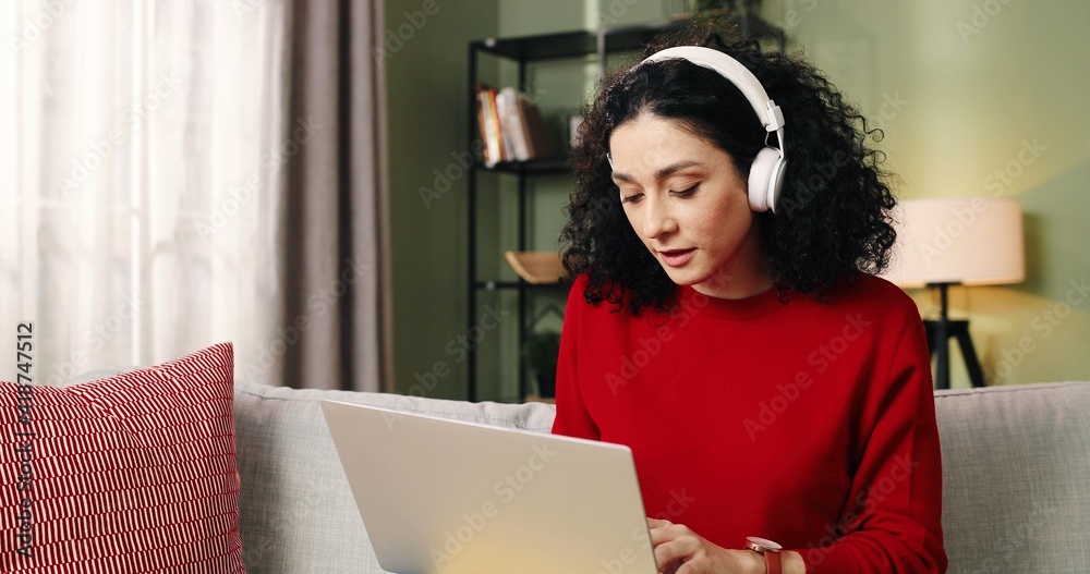 Portrait of happy Caucasian pretty young woman sitting on sofa in living room listening to song in headphones while working typing on laptop searching internet, freelance works at home. Job concept