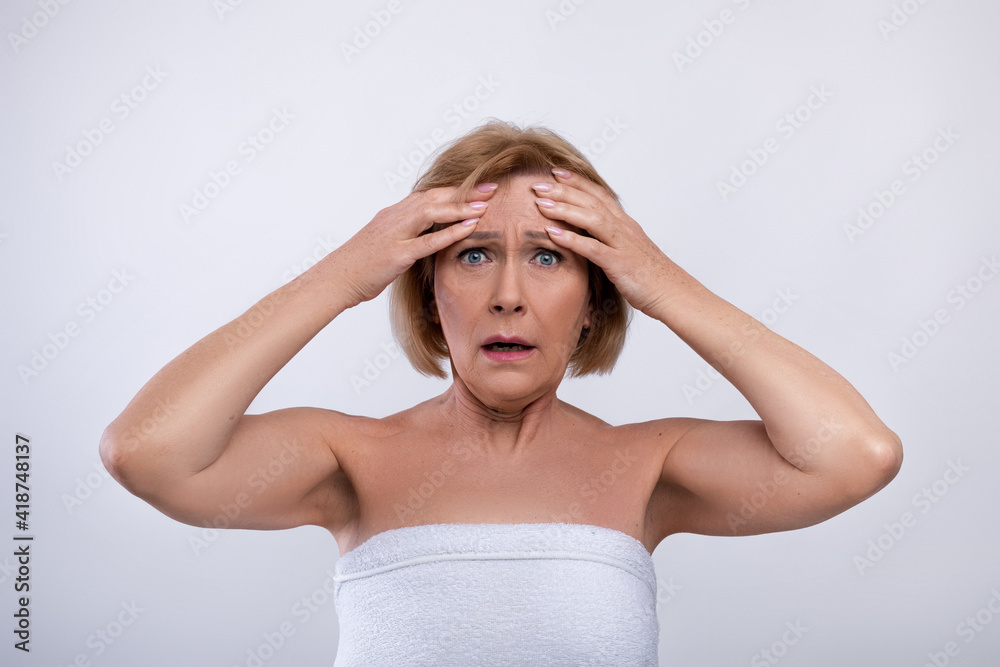 Portrait of terrified senior lady touching her forehead, scared to see new wrinkles on light studio background