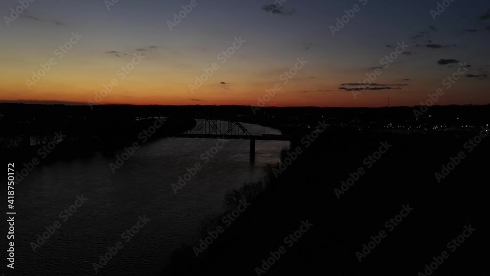 Sunset with amazing colors arch steel bridge over a river with black silhouettes of trucks traveling over and city lights on the side on a bright day in the USA Slowly panning drone shot Sunset with amazing colors arch steel bridge over a river with black silhouettes of trucks traveling over and city lights on the side on a bright day in the USA Slowly panning drone shot