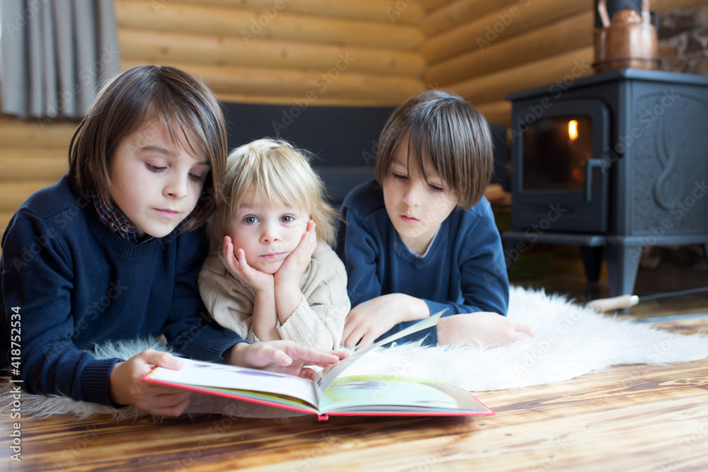 Three sweet children, siblings lying on the floor in little fancy wooden cottage, reading a book, drinking tea Three sweet children, siblings lying on the floor in little fancy wooden cottage, reading a book, drinking tea