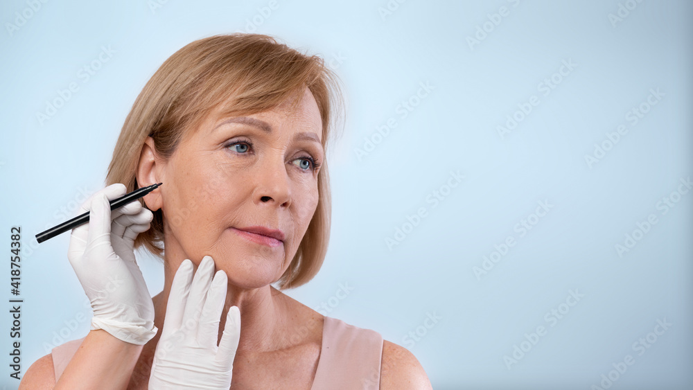 Plastic surgeon drawing lines on mature woman's face for cosmetic operation on blue studio background, banner design Plastic surgeon drawing lines on mature woman's face for cosmetic operation on blue studio background, banner design