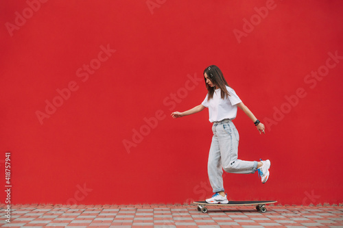 Full-length shot of a skater woman wearing fancy clothes and riding her skateboard in front of a red wall with a copy space on a city street.