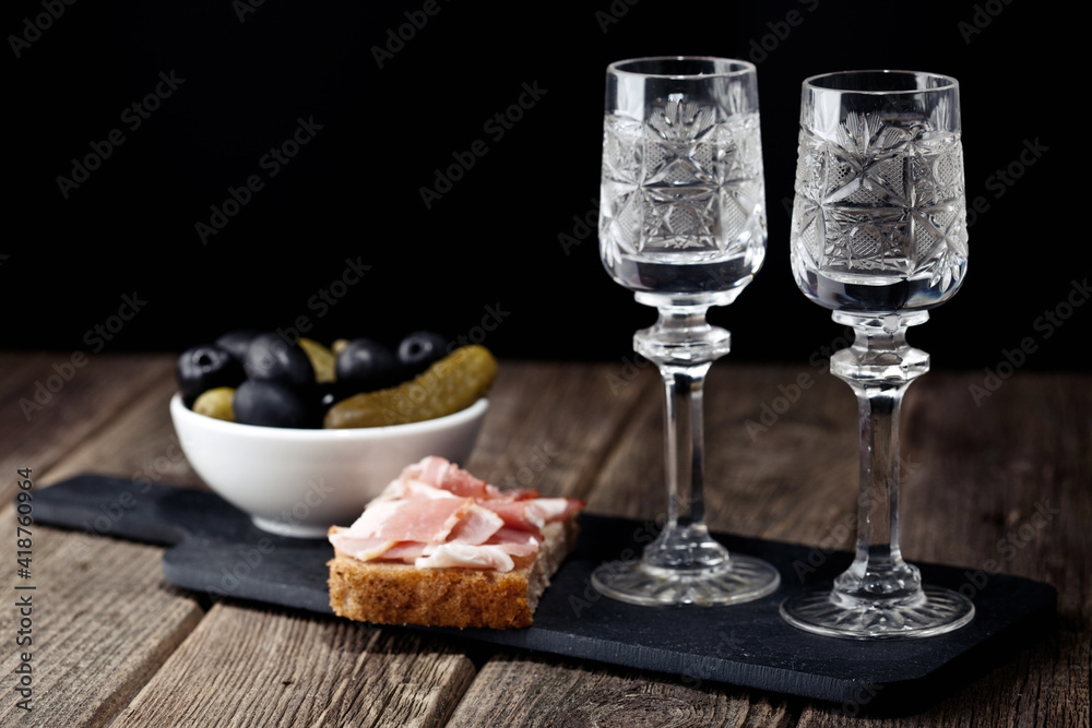 vodka in glasses on a black shale snack tray black bread with bacon pickled olive cucumbers on a wooden table vodka in glasses on a black shale snack tray black bread with bacon pickled olive cucumbers on a wooden table