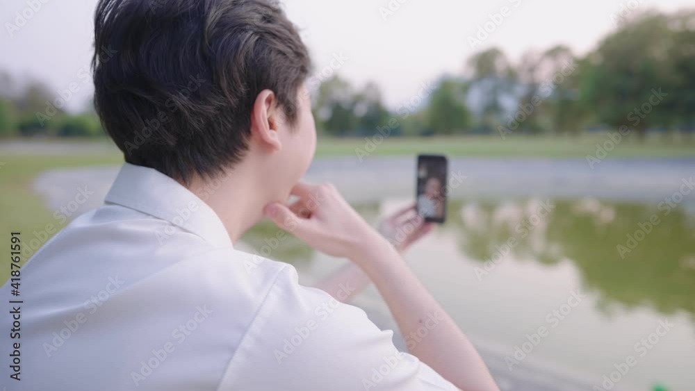 Happy young man holding a smartphone, looking at the screen, talking on a video call while sitting on the grass in the park, online technology, mobile application, social distancing, self isolation Happy young man holding a smartphone, looking at the screen, talking on a video call while sitting on the grass in the park, online technology, mobile application, social distancing, self isolation