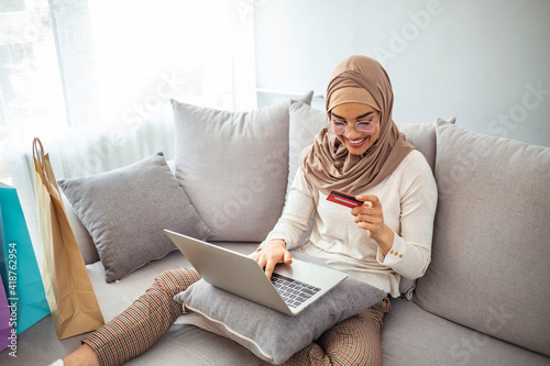 Close up shot of Muslim mother buying groceries online from her laptop at home, paying with credit card, during Coronavirus pandemic. Shot of a young woman using a laptop and credit card at home © Dragana Gordic