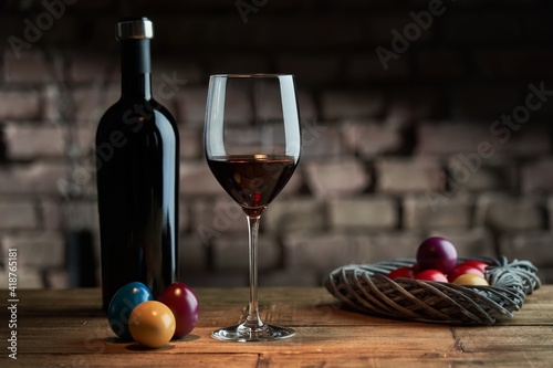Easter decorations, colorful eggs, a glass of red wine with a bottle on table at home dark warm living room, brick wall in background. Copy space for text.