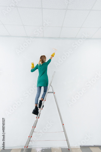 A young girl on a stepladder paints a white wall with a roller. Repair of the interior.