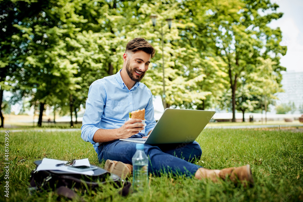 Happy young man in park using laptop eating sandwich Happy young man in park using laptop eating sandwich