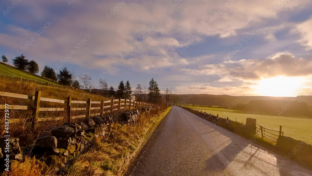 A moody image taken along a rural road with a wooden fence and drystone wall leading into the distance. All lit by a low sun with soft clouds and blue sky.