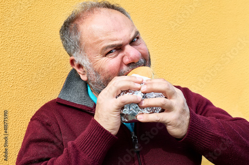 Funny bearded mature man eating sandwich on the street against a yellow wall. Man hungry like a wolf biting bread. Unhealthy eating.