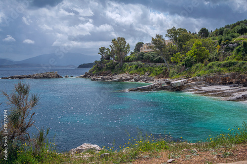 Beautiful landscape - sea lagoon with turquoise calm water, stones and rocks on the beach, clouds on the sky, green trees and bushes, mountains on the horizon. Corfu Island, Greece, Kassiopi village