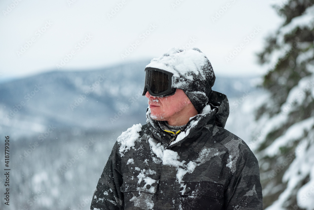 Snowboarder man with snow covered face is standing on the mountains background.