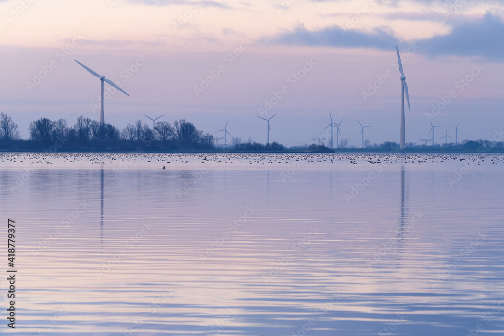 Lake with two Bladed Wind Turbines, or modern Windmills, in a Wind Farm along the nature reserve Eemmeer in the Netherlands. Cold evening colours with waterfowl on a lake.