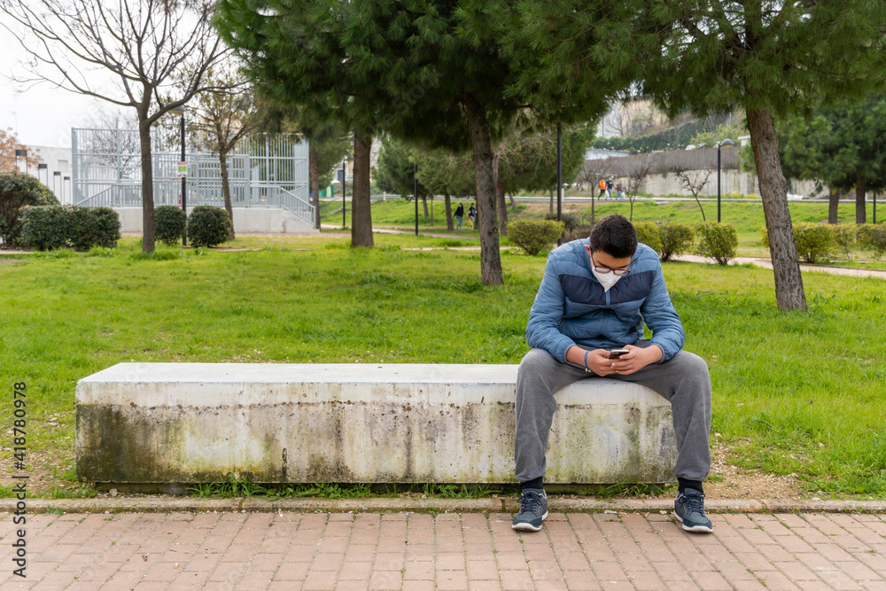 Fototapeta premium Young boy wearing face mask due to covid19 using his cellphone while sitting in a bench in the park.